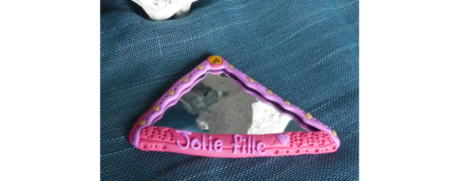 Un miroir triangulaire, rose, avec l'inscription "jolie fille", des éléments décoratifs en relief et des perles.